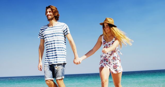 A young Caucasian couple enjoys a leisurely walk along a sunny beach, with copy space. Their casual attire and joyful expressions suggest a carefree, romantic moment shared between them.