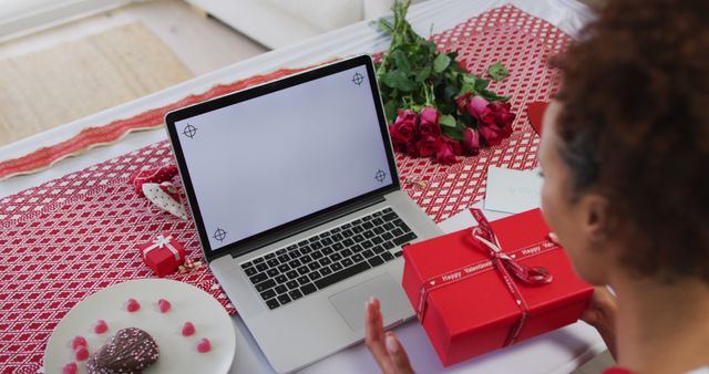 Ideal for articles and media on Valentine's Day celebrations, online gifting, and long-distance relationships. Can be used for promoting online gift stores, romantic content, or Valentine's Day specials. The setting highlights romance with a decorated red tablecloth, roses, a laptop hinting at an online connection, and a festive gift box.