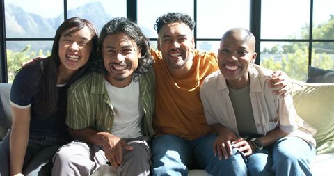 Smiling diverse friends sitting on sofa in sunlit living room with mountain view