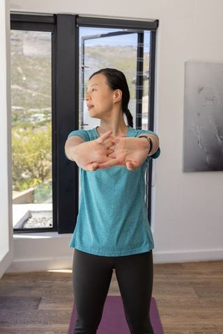 Asian woman stretching at home by window on yoga mat for morning fitness and wellness