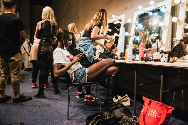 Group of models and stylists preparing for a fashion show backstage. Models are sitting and using their phones while stylists are doing hair and makeup. Ideal for depicting the behind-the-scenes hustle of the fashion industry, makeup artistry, and the energy of fashion events.