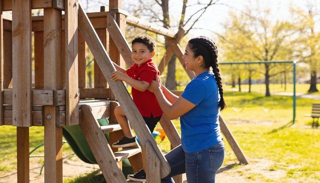 Caregiver steadying young boy climbing wooden play ladder in sunny park, smiling child