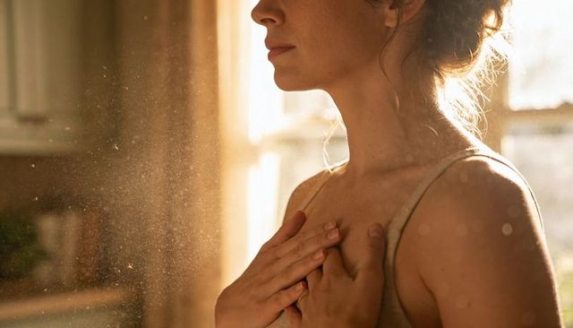 Woman standing by sunlit window placing hands on chest, backlit dust motes, serenity