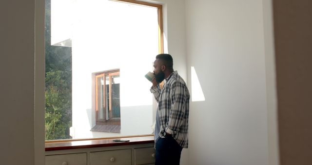 A man is drinking coffee while standing near a window in a bright and modern home. This image can be used for home living articles, lifestyle blogs, relaxation techniques, interior design inspiration, or marketing for modern housing.