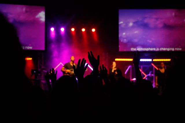 This image depicts a lively concert audience with hands raised high, immersed in the music and atmospheric lighting. Ideal for promoting live music events, spiritual gatherings, music festivals, and performing arts advertisements. Can be used in blogs, articles, and websites about live music experience, crowd engagement, and concert atmosphere.