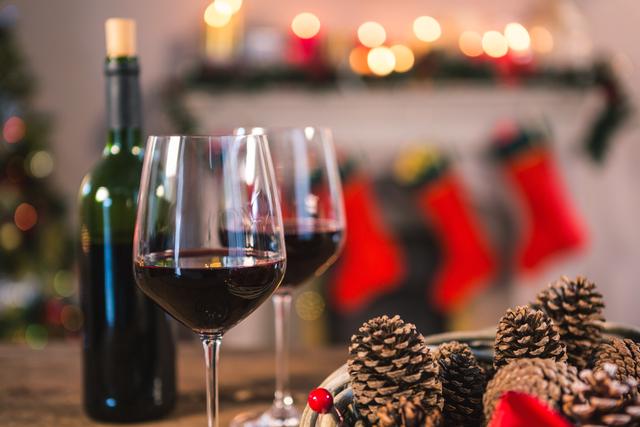 Red wine bottle and glasses on wooden table with pine cones, in front of a decorated fireplace with Christmas stockings. Ideal for holiday marketing, festive greeting cards, seasonal blog posts, and home decor inspiration.