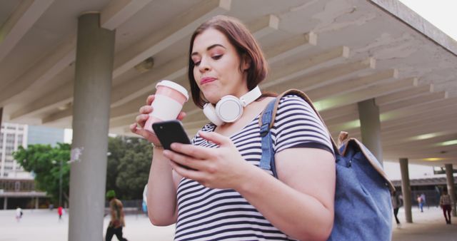 A young woman wearing a striped shirt is drinking coffee while using her smartphone outdoors. She has headphones around her neck and carries a backpack. The urban setting in the background includes concrete pillars and trees, enhancing the casual and modern atmosphere of the scene. This can be used for themes related to lifestyle, technology, city life, and youth activity.