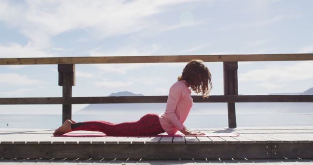 Woman practicing cobra pose on yoga mat during outdoor yoga session with ocean and mountains in background. Ideal for content promoting healthy lifestyle, mindfulness, relaxation, outdoor activities, and travel destinations. Useful for fitness blogs, wellness programs, yoga tutorials, and vacation advertisements.