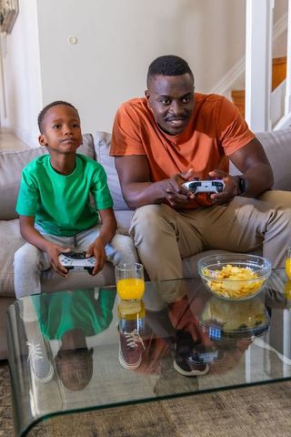 African American father and son playing video games on sofa, bonding with controllers