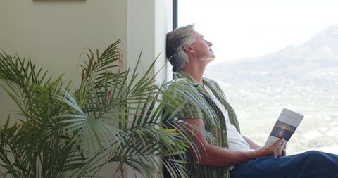 Mature man relaxing by window, reading brochure beside indoor palm and scenic valley view