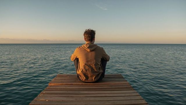 Solitary man sitting on wooden pier gazing at ocean horizon during golden hour