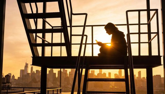 Silhouetted person sitting on fire escape at sunset holding smartphone over city skyline