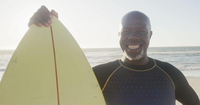 Smiling senior man wearing wetsuit holding surfboard at beach with ocean backdrop. Perfect for projects on active aging, retirement lifestyles, outdoor activities, and summer fun.
