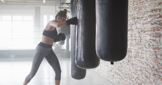 A dedicated woman is engrossed in an intense boxing workout, focusing her punches on a heavy bag inside an industrial-style gym. This visual is ideal for illustrating themes around women’s fitness, athletic training, strength path ways, and sports motivation. It may be used in promotional materials for fitness programs, gyms, sportswear advertisements, or health-related websites.