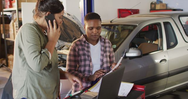 Image of two diverse female car mechanics using laptop and smartphone, searching for car parts. working in car repair shop and running small feminine business concept.