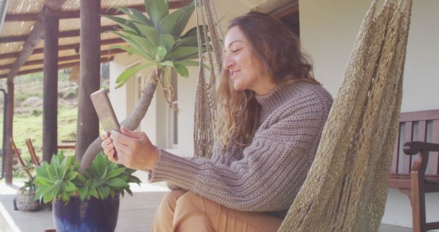 Smiling woman sits comfortably in a hammock on a patio while using her smartphone. She is wearing a cozy knit sweater and casual attire, creating a relaxed and serene ambiance. This image is perfect for promoting digital communication, outdoor leisure activities, or lifestyle content emphasizing relaxation and connectivity with nature.