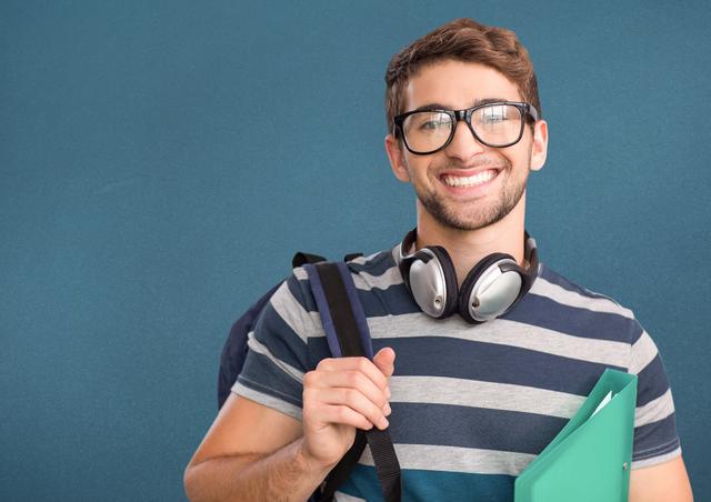 Young male college student smiling confidently while holding a file and wearing headphones around his neck. He is carrying a backpack and dressed in a casual striped shirt. Ideal for use in educational materials, school promotions, student lifestyle blogs, and academic advertisements.