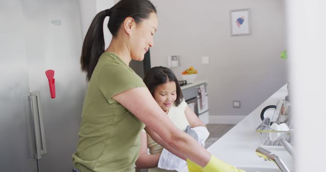 Mother wearing rubber gloves is joyfully helping her daughter wash dishes in a modern kitchen. The image showcases family bonding, teamwork, and involvement in household chores. Suitable for use in articles or advertisements related to family life, household products, cleaning services, or parenting tips.