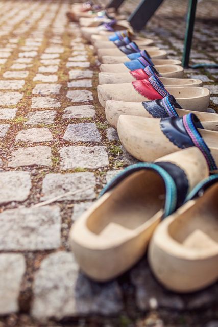 Wooden clogs neatly lined up on a cobblestone pathway under warm sunlight. The presence of colorful straps contrasts with the natural wood, creating a picturesque scene that evokes traditional handicrafts and cultural heritage. Ideal for use in blogs, articles focusing on traditional crafts, travel advertisements, and cultural heritage sites.