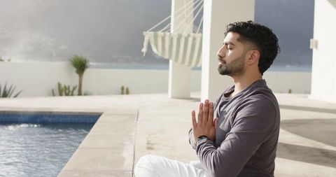 Indian man practicing mindfulness meditation by pool on coastal terrace with sea view