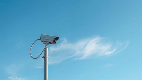 Surveillance camera monitoring urban perimeter from tall metal pole against blue sky