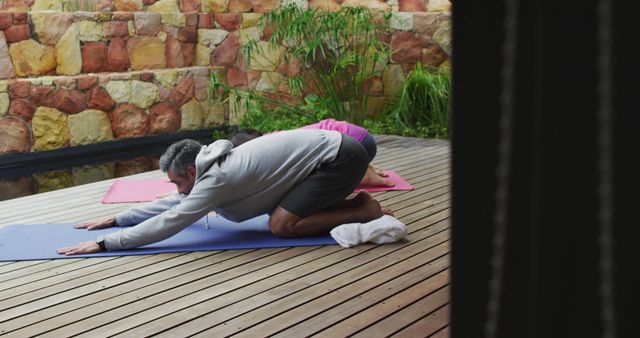 Couple practicing yoga on colorful mats on a wooden deck, surrounded by greenery. The man is performing the child's pose, focusing on relaxation and stretching. The outdoor environment adds to the calm and serene atmosphere. Perfect for promotions of wellness retreats, yoga classes, and outdoor exercise programs.