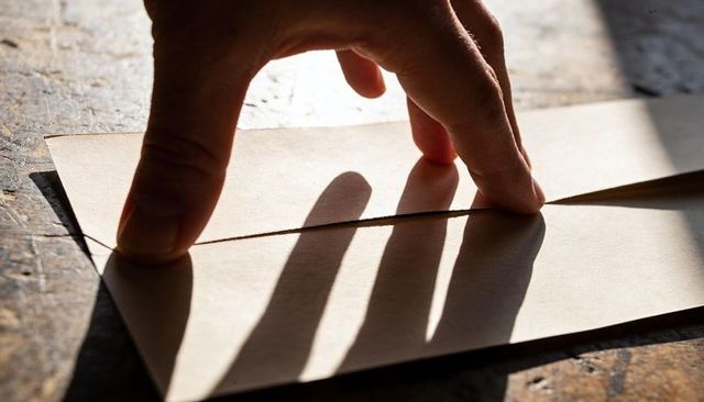 Pressing fingers closing brown kraft envelope on sunlit wooden desk casting long shadows