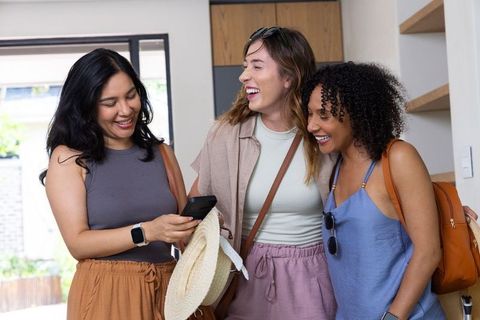 Multicultural female friends laughing while sharing smartphone in sunlit entryway