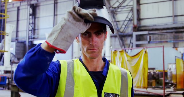 Man wearing high-visibility vest and gloves, lifting welding helmet in an industrial factory environment. This image can be used for advertisements related to industrial safety standards, manufacturing processes, engineering services, and employment in manufacturing sectors.