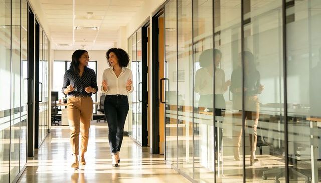 Two colleagues walking and chatting through sunlit office corridor with glass reflections