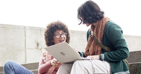 Diverse female students sharing laptop on campus steps, collaborating and studying outdoors
