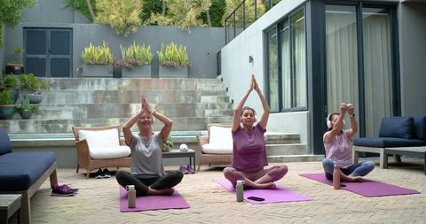 Diverse women practicing seated yoga on sunny patio with purple mats, water bottles and cushions