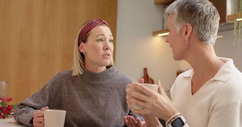 Women chatting over coffee at cozy kitchen counter wearing sweaters and smartwatch