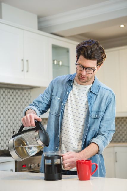 Man in casual attire pouring coffee into a French press in a modern kitchen. Ideal for use in content related to morning routines, coffee preparation, home life, kitchen appliances, and casual lifestyle. Suitable for blogs, advertisements, and social media posts about coffee culture and domestic activities.