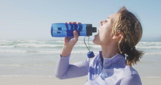 Caucasian woman wearing sports clothes holding water bottle and drinking at beach. Sport, healthy and active lifestyle.
