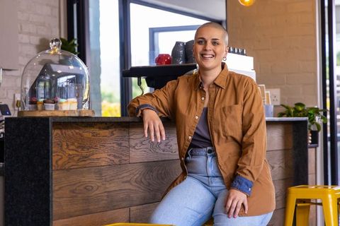Smiling barista sitting on yellow stool at cozy coffee counter with pastry dome