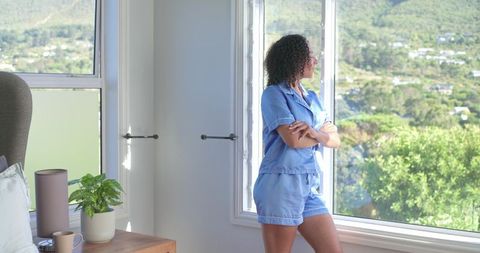 African woman standing at window in blue pajamas enjoying sunlit bedroom morning view