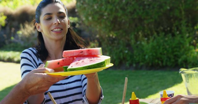 A young Caucasian woman is offering slices of watermelon at an outdoor gathering, with copy space. Her cheerful expression and the casual setting suggest a relaxed and enjoyable social event.