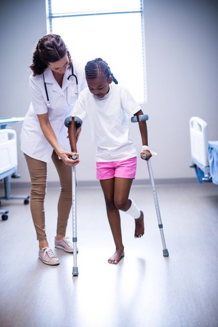 Female doctor assisting girl to walk with crutches in ward of hospital