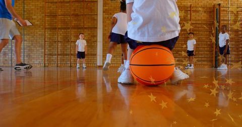 Crouching boy holding basketball on school gym floor during primary pe class with coach