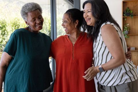 African american senior women laughing and hugging in sunlit home, showing smartwatch