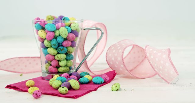 Multicolored speckled eggs are displayed in a clear glass cup, accompanied by a pink ribbon and placed on a vibrant pink cloth. Perfect for use in Easter-related designs, holiday card creation, festive home decor inspiration, or seasonal advertising campaigns. Bright pastel colors make the scene cheerful and ideal for spring-themed projects.