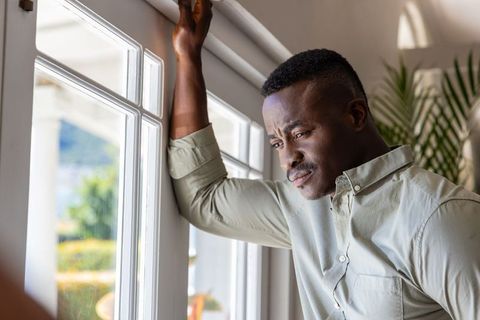 Contemplative african american man leaning on window frame reflecting in sunlit interior