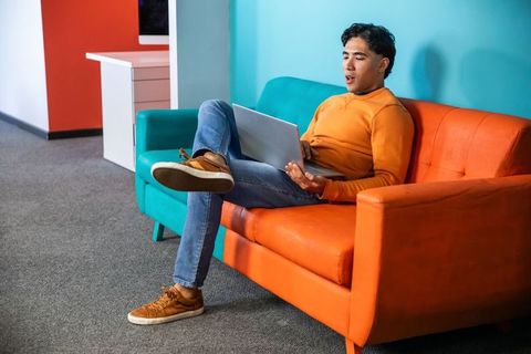 Young asian man working on laptop on colorful orange couch in modern coworking lounge