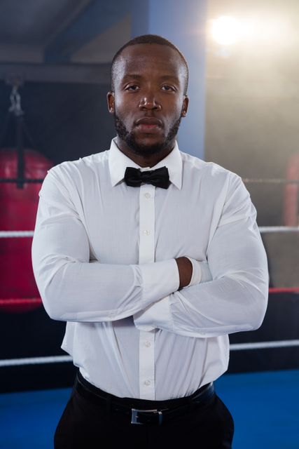 Young male referee standing with arms crossed in boxing ring, exuding confidence and authority. Ideal for use in sports-related articles, promotional materials for boxing events, or advertisements highlighting professionalism and athleticism.