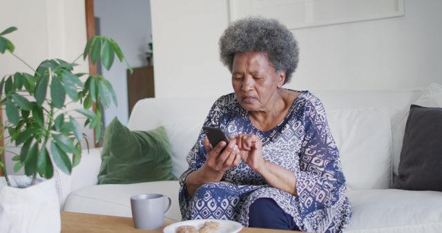 Elderly woman wearing casual clothes sitting on a couch, using her smartphone with a focused expression. Ideal for depicting seniors' engagement with technology, leisure time activities, or modern lifestyle of retired people. Can be used in advertisements for tech products, senior living, or healthcare services.