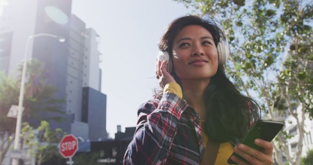 Young woman in casual clothing, smiling and listening to music on her headphones while holding a smartphone. She is outside in an urban environment, suggested by the outdoor scenery and city buildings. This image is perfect for promoting music streaming services, lifestyle blogs, technology products, or urban living themes.