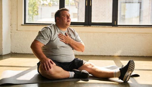 Overweight man pausing workout on yoga mat in sunlit studio showing breath recovery