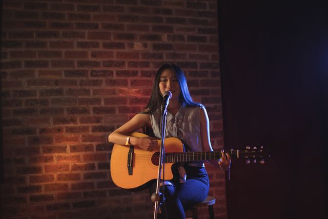 Young female singer playing guitar and singing on stage in a nightclub. Ideal for use in articles about live music, entertainment, young musicians, and nightlife. Suitable for promoting music events, concerts, and acoustic performances.