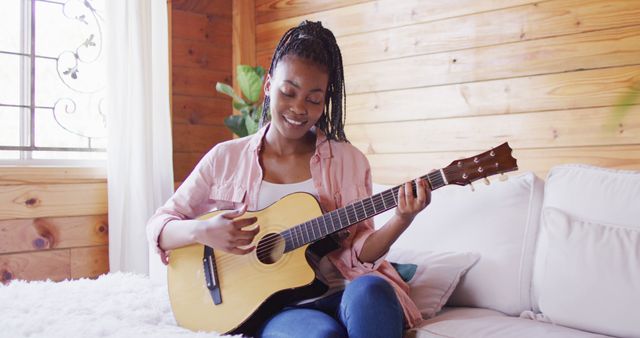 Young woman happily playing an acoustic guitar inside a cozy, wooden house. Perfect for use in ads and articles about music, hobbies, relaxation, home lifestyles, cultural diversity, and leisure activities.